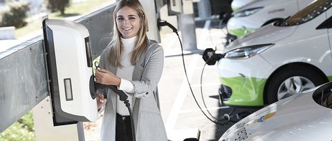 Eine Frau mit Blazer lacht in die Kamera und löst gerade einen Ladevorgang an einer Ladestation aus. Im Hintergrund ist eine Flotte von Elektrofahrzeugen und weitere Wallboxen zu sehen 