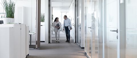 Zwei Energiedienst-Mitarbeiter unterhalten sich während sie laufen im Büro in Rheinfelden. Das Büro ist hell und es ist eine grüne Pflanze zu sehen.
