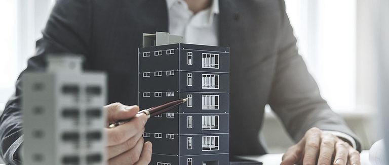 Ein Geschäftsmann im Anzug zeigt mit einem Kugelschreiber auf ein Modell eines Mehrfamilienhauses