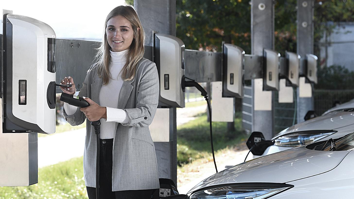 Eine Frau mit Blazer lacht in die Kamera und löst gerade einen Ladevorgang an einer Ladestation aus. Im Hintergrund ist eine Flotte von Elektrofahrzeugen und weitere Wallboxen zu sehen 