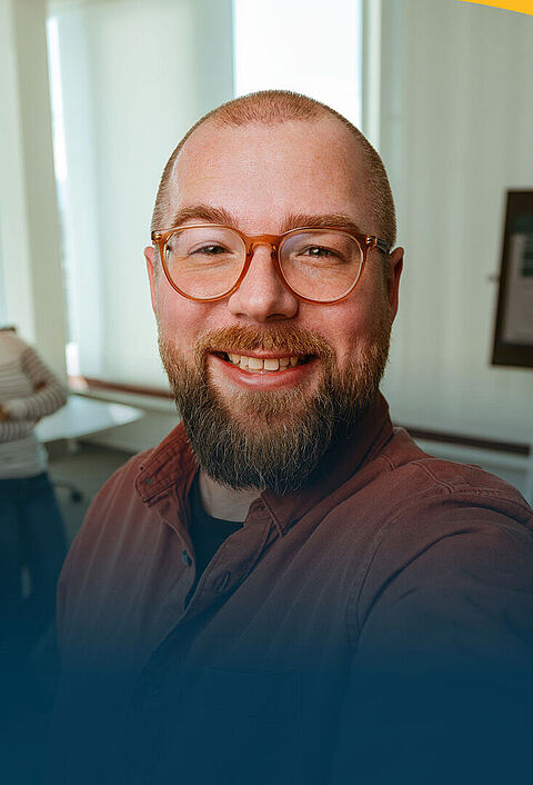 Ein lächelnder Mann mit Brille macht ein Selfie im Büro. Im Hintergrund sieht man drei seiner Kollegen bei der Arbeit.