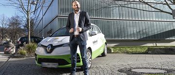Nils Hösch, Leiter des Bereichs NaturEnergie Elektromobilität bei Energiedienst steht vor einem Elektroauto von my-e-car, welches vor einem Galsgebäude steht.