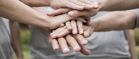 Hände eines mehrköpfigen Teams mit heller Kleidung liegen aufeinander. Teamzusammenhalt soll ausgedrückt werden.