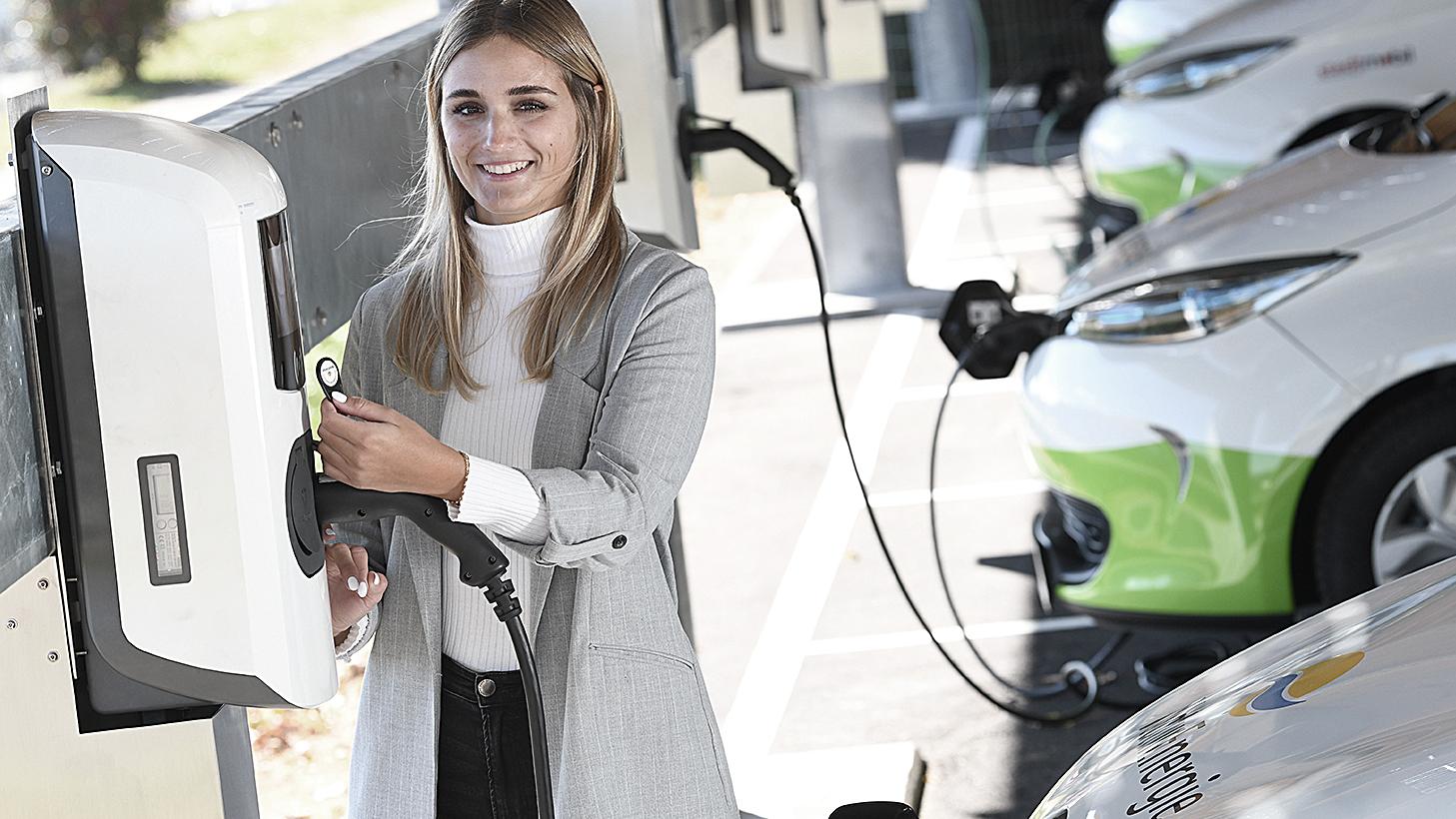 Eine Frau mit Blazer lacht in die Kamera und löst gerade einen Ladevorgang an einer Ladestation aus. Im Hintergrund ist eine Flotte von Elektrofahrzeugen und weitere Wallboxen zu sehen 