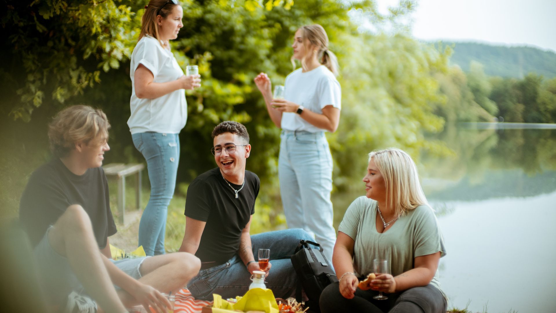 Das Bild zeigt mehrere Menschen bei einem Picknick. Im Hintergrund ist ein See zu sehen. Zwei Menschen stehen, drei sitzen auf einer Decke auf dem Boden und unterhalten sich. 