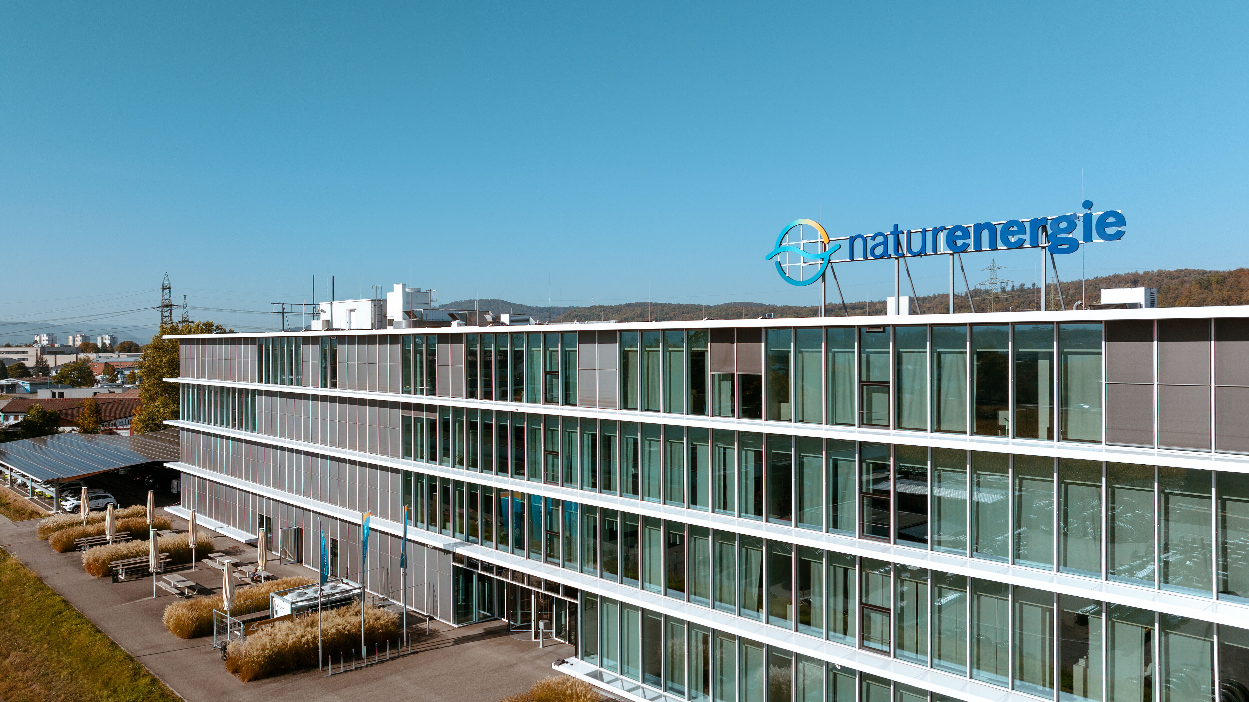 Außenaufnahme eines Bürogebäudes. Das Gebäude hat eine große Glasfront. Vor dem Gebäude wehen naturenergie-Fahnen. Es gibt einen Außensitzplatz mit Bänken und Sonnenschirmen.
