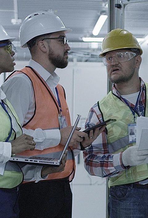 Gruppe multiethnischer Männer und Frauen in Hardhats, die in der Halle des Solaranlagenkontrollzentrums tätig sind, diskutiert zwischen Racks