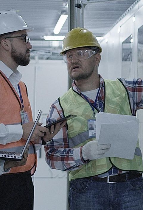 Gruppe multiethnischer Männer und Frauen in Hardhats, die in der Halle des Solaranlagenkontrollzentrums tätig sind, diskutiert zwischen Racks