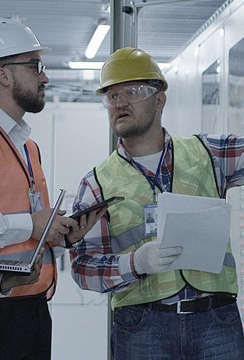 Gruppe multiethnischer Männer und Frauen in Hardhats, die in der Halle des Solaranlagenkontrollzentrums tätig sind, diskutiert zwischen Racks