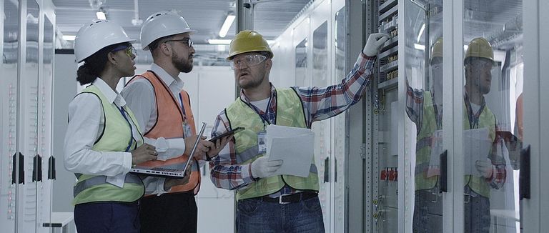 Gruppe multiethnischer Männer und Frauen in Hardhats, die in der Halle des Solaranlagenkontrollzentrums tätig sind, diskutiert zwischen Racks