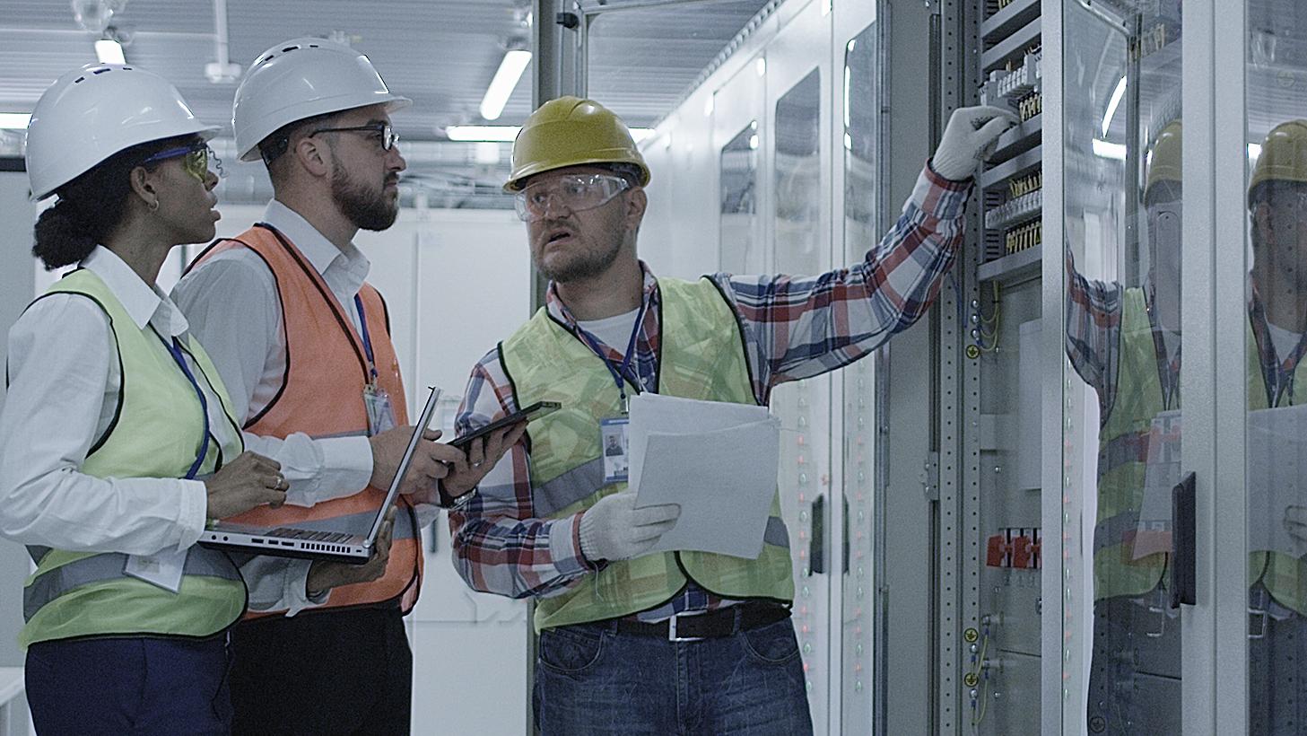 Gruppe multiethnischer Männer und Frauen in Hardhats, die in der Halle des Solaranlagenkontrollzentrums tätig sind, diskutiert zwischen Racks