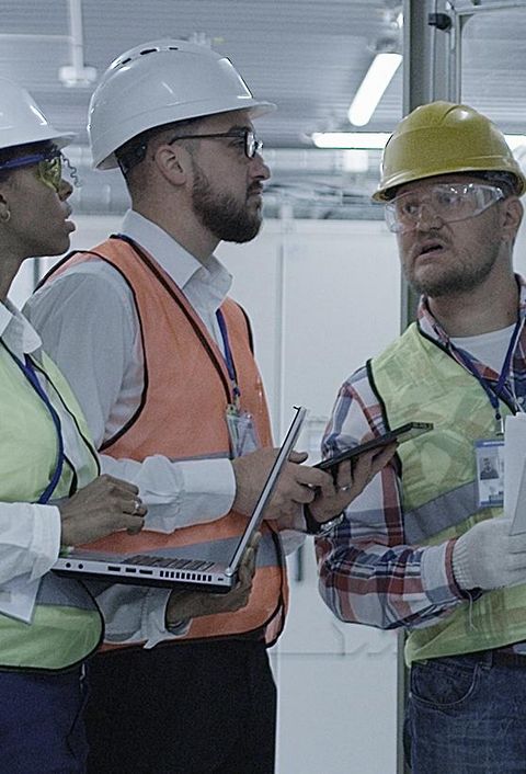 Gruppe multiethnischer Männer und Frauen in Hardhats, die in der Halle des Solaranlagenkontrollzentrums tätig sind, diskutiert zwischen Racks