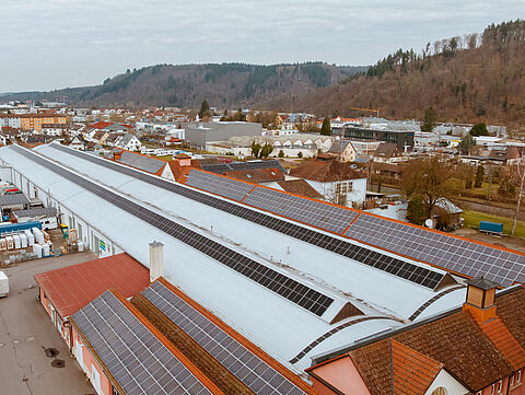 Luftaufnahme von Solardächern im Industriegebiet Großes Industriegebäude mit Solarpaneelen auf dem Dach, umgeben von Fahrzeugen, Containern und Wohnwagen im Hintergrund.
