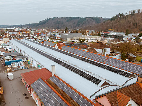 Großes Industriegebäude mit Solarpaneelen auf dem Dach, umgeben von Fahrzeugen, Containern und Wohnwagen im Hintergrund.