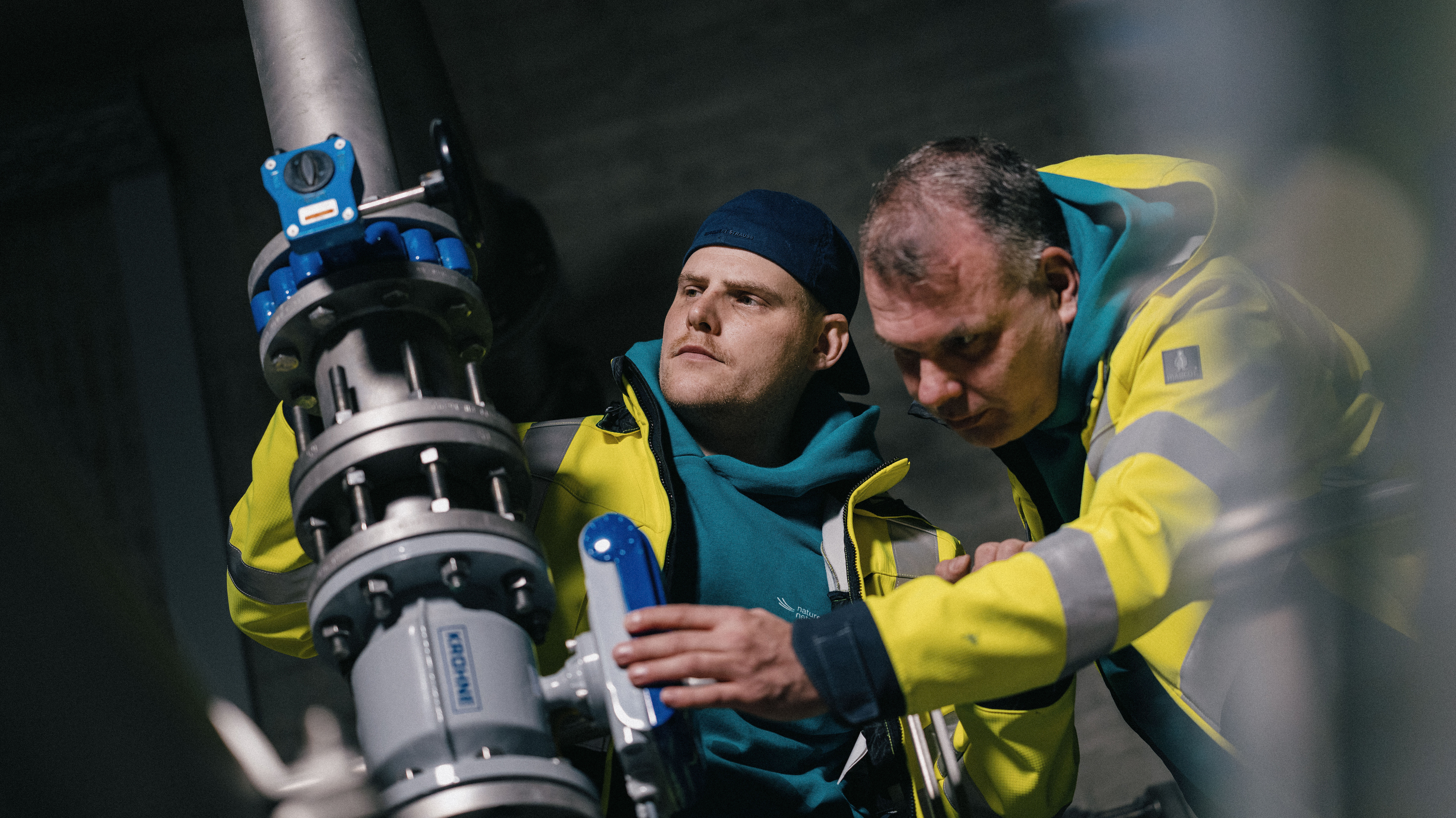 Zwei Techniker in Schutzkleidung arbeiten gemeinsam an einer großen Rohrleitung und prüfen ein Ventil.