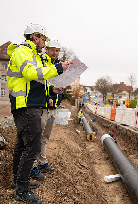 Zwei Personen mit Helmen prüfen Pläne an einer Baustelle mit offenem Rohrgraben.