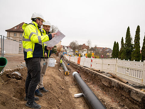 Zwei Personen mit Helmen prüfen Pläne an einer Baustelle mit offenem Rohrgraben.