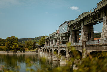 Ein Wasserkraftwerk von naturenergie, das sich über einen Fluss erstreckt. Die Struktur des Kraftwerks besteht aus mehreren Steinpfeilern und Metallbrücken. Im Hintergrund sind grüne Bäume und Hügel zu sehen