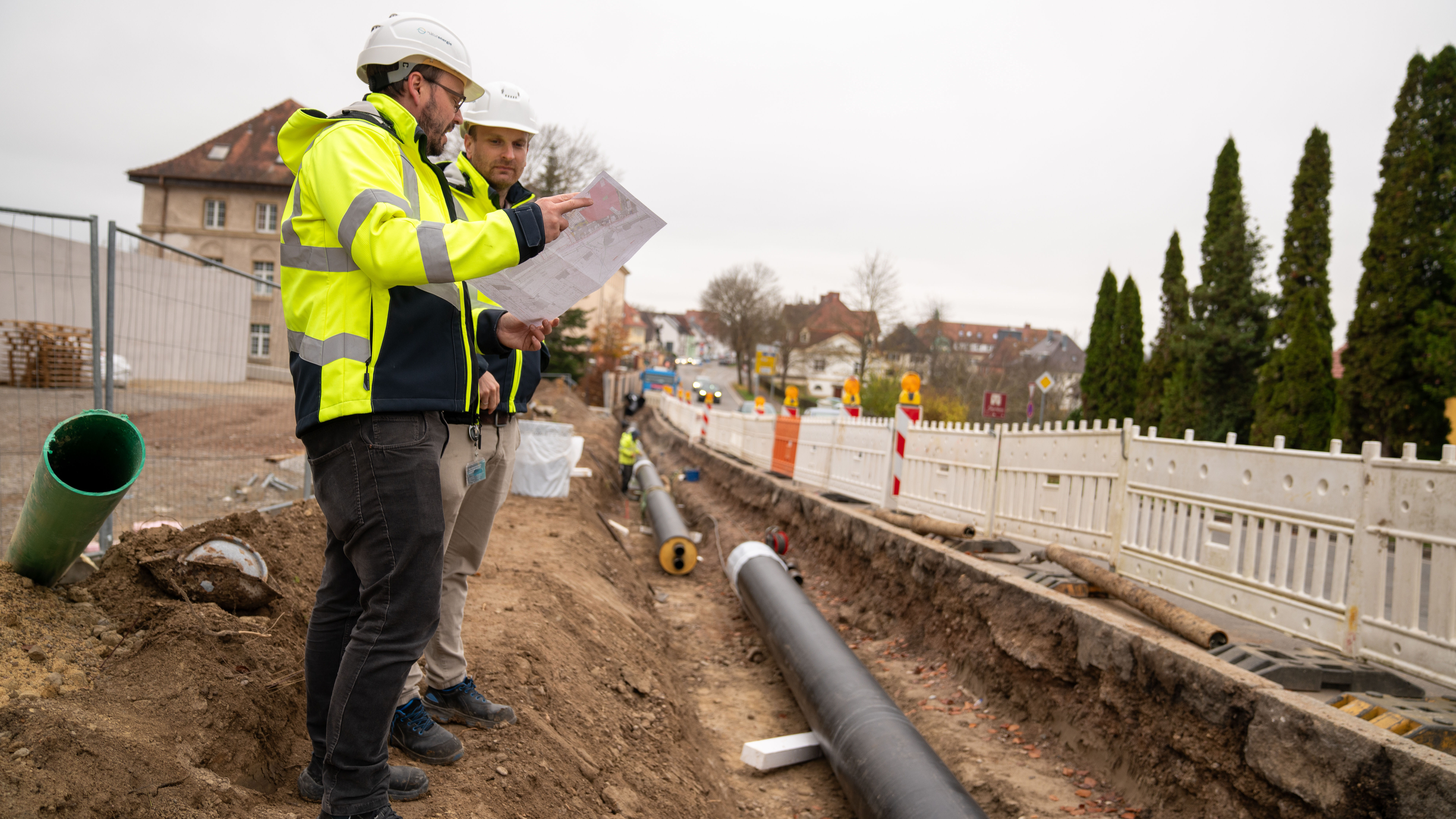 Zwei Bauingenieure mit Helmen besprechen Baupläne an einer Baustelle mit Rohrleitung im Graben.