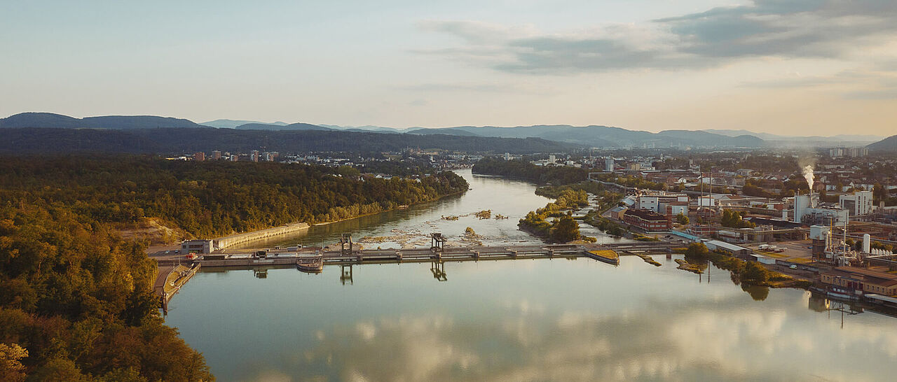 Luftaufnahme eines Flusses mit einem Wasserkraftwerk und einer Stadt im Hintergrund.