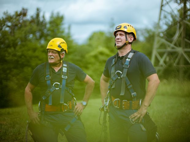 Das Bild zeigt zwei Menschen in Kletterausrüstung und Schutzhelmen. Im Hintergrund ist ein Strommast zu sehen. 