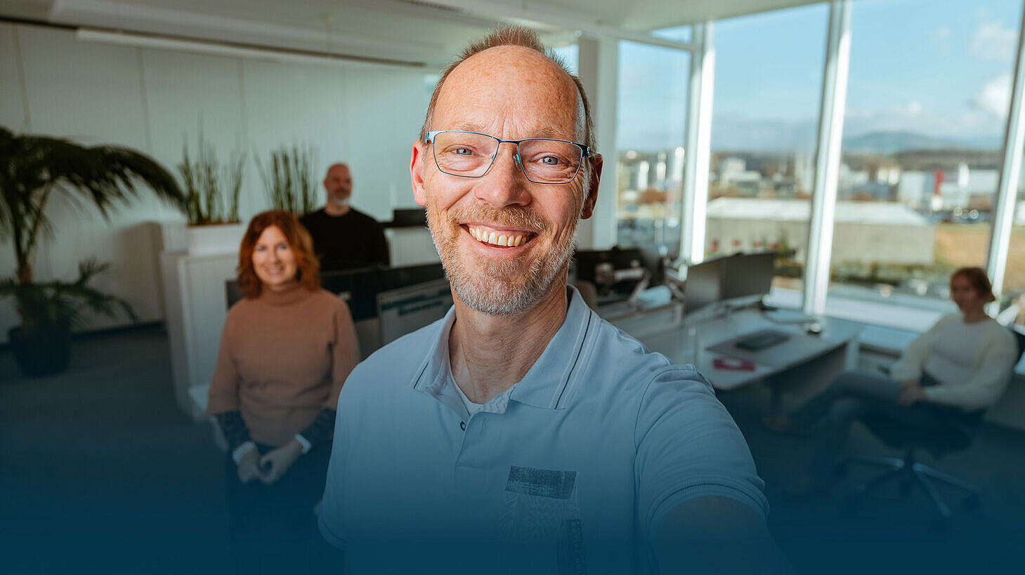Ein älterer Mitarbeiter macht ein Selfie im Büro. Im Hintergrund sieht man drei seiner Kollegen an ihren Arbeitsplätzen.