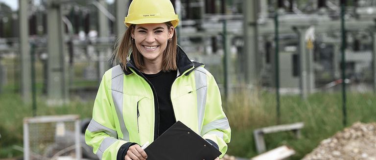 Nahwärme-Ingenieurin von NaturEnergie in Arbeitsschutzkleidung lächelt in die Kamera und hält ein Schreibbrett in der Hand.