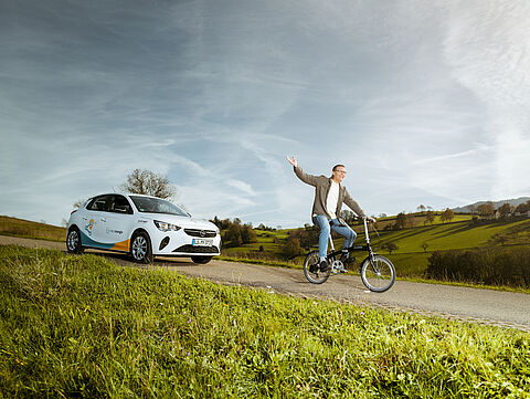 Mann auf Fahrrad und dahinter Carsharing Auto von naturenergie