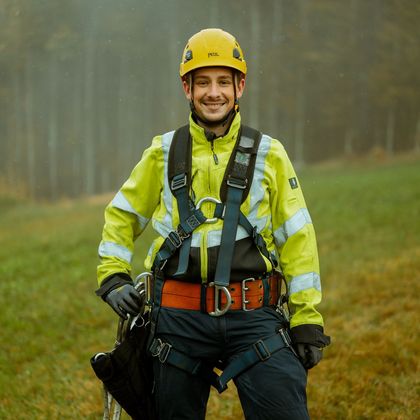 Das Bild zeigt eine Person welche Arbeitskleidung und Schutzhelm trägt und in der Natur auf einer Grünfläche steht 