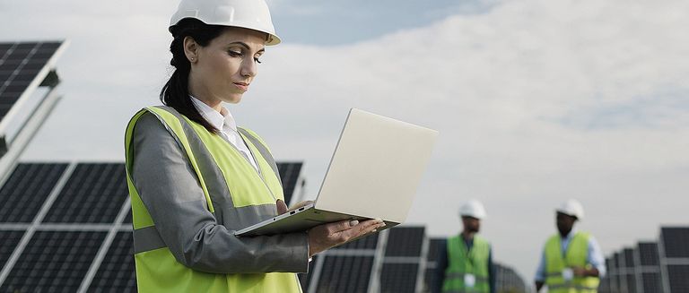 Elektroingenieurin in Schutzhelm und Uniform mit Laptop prüft Solarpaneele. 