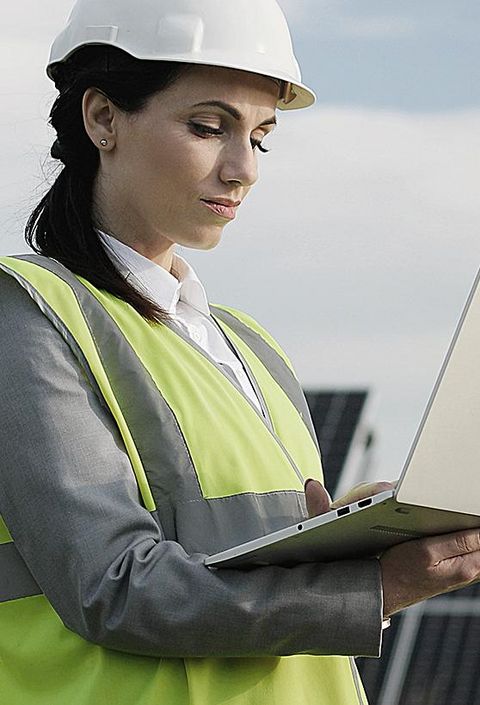 Elektroingenieurin in Schutzhelm und Uniform mit Laptop prüft Solarpaneele. 