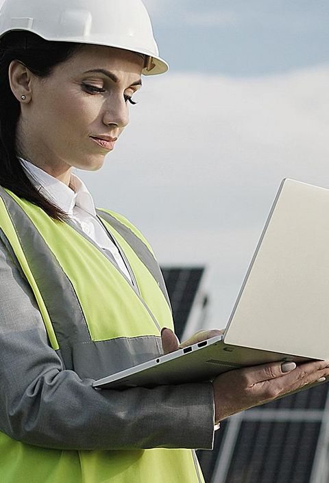 Elektroingenieurin in Schutzhelm und Uniform mit Laptop prüft Solarpaneele. 