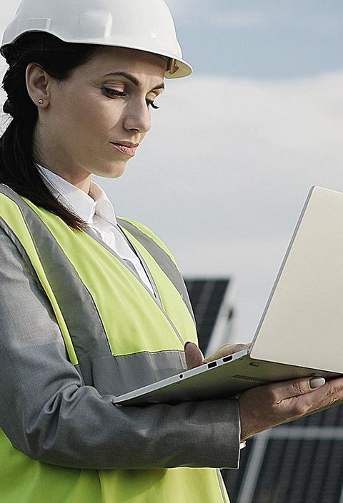 Elektroingenieurin in Schutzhelm und Uniform mit Laptop prüft Solarpaneele. 
