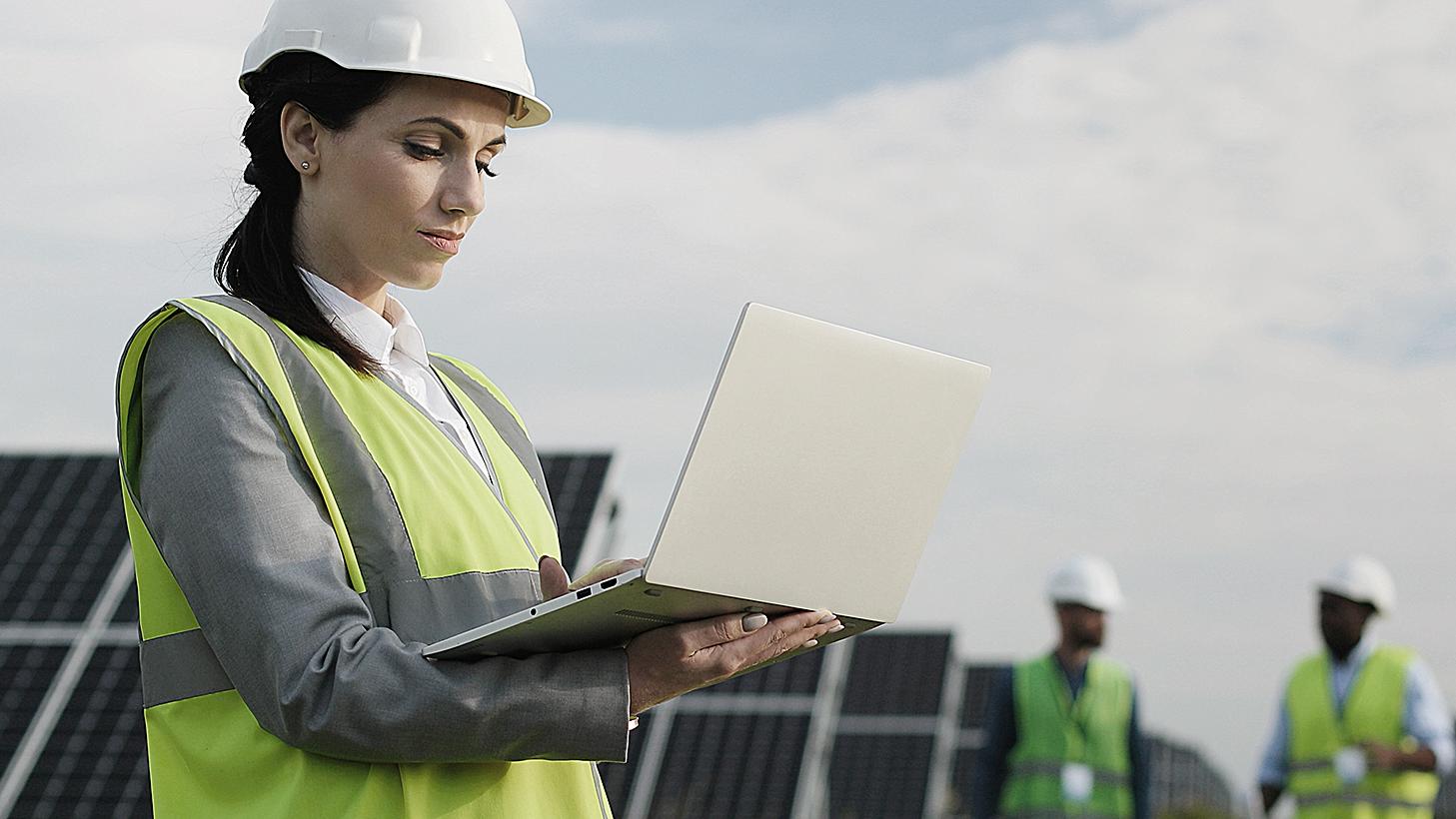 Elektroingenieurin in Schutzhelm und Uniform mit Laptop prüft Solarpaneele. 