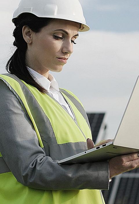 Elektroingenieurin in Schutzhelm und Uniform mit Laptop prüft Solarpaneele. 