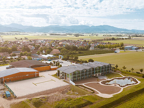 Luftaufnahme mit modernen Gebäuden, Feldern und Dorf im Hintergrund