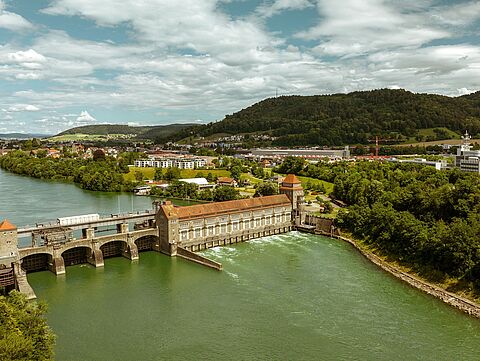 Fluss mit Staudamm, roten Dächern und Stadt im Hintergrund