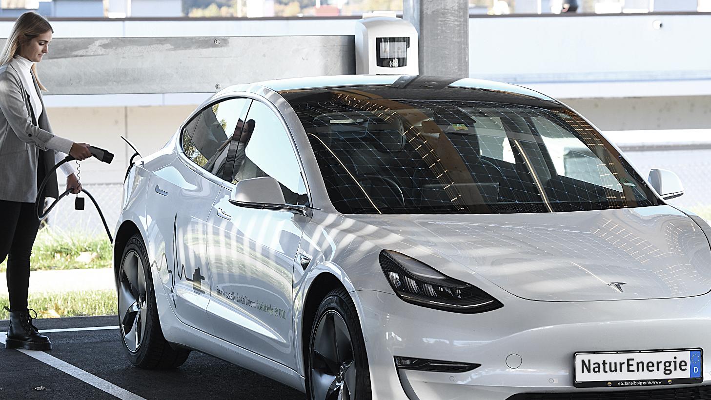 Frau bewegt sich mit Ladestecker zu einem weißen Elektroauto von Tesla, um  es mit Strom zu tanken