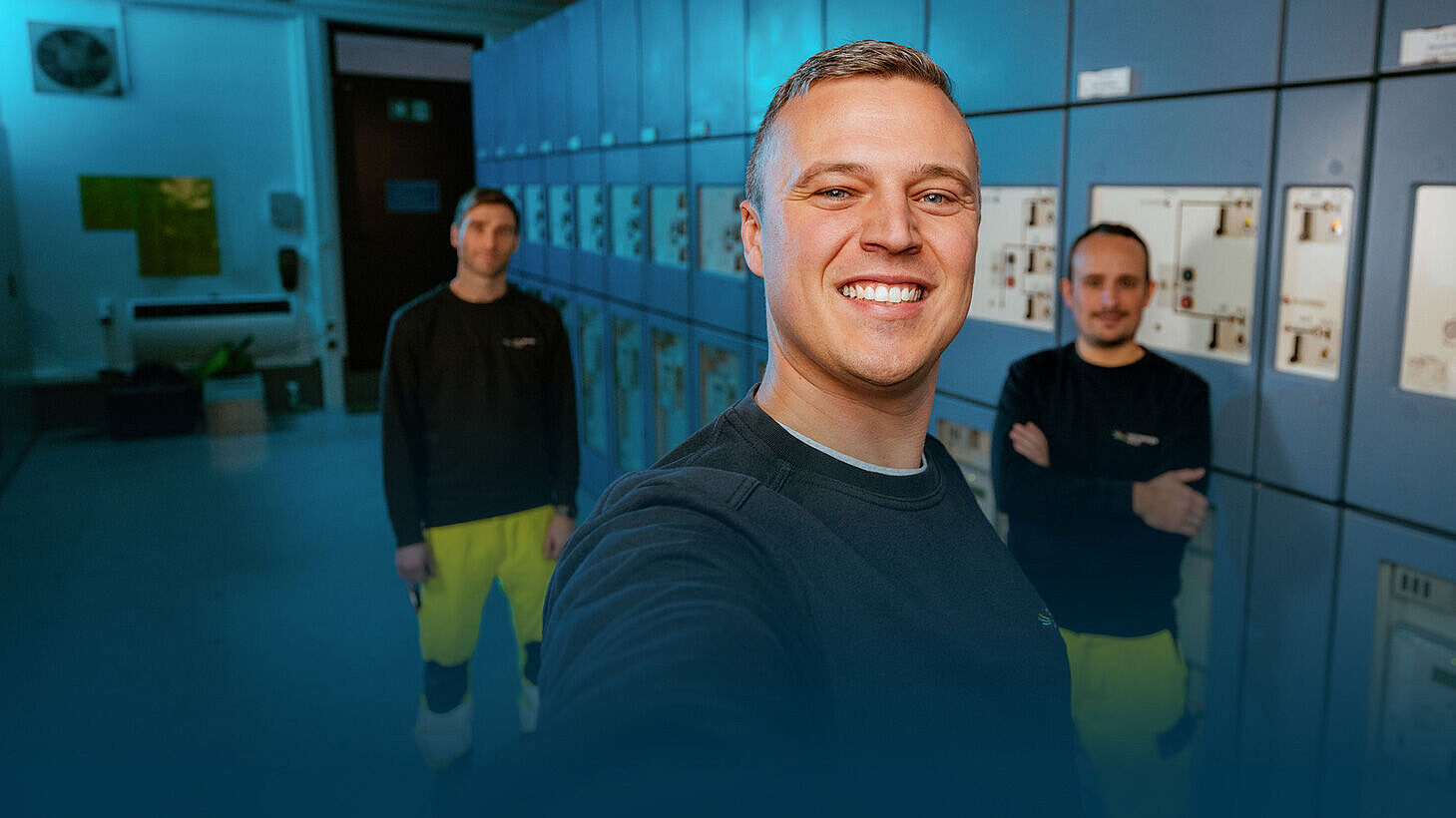 Ein Mann macht ein Selfie in einem technischen Raum mit Schaltschränken. Hinter ihm sind zwei seiner Kollegen zu sehen.