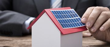 Geschäftsmann legt Papierbild einer Solaranlage auf ein kleines Hausmodell vor sich auf dem Tisch