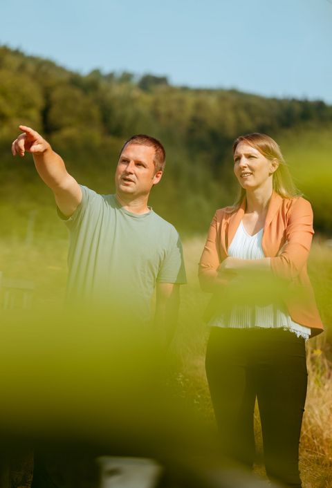 Das Bild zeigt zwei Menschen in der Natur. Eine Person zeigt der anderen Person gerade etwas und zeigt mit der Hand darauf. Im Vordergrund ist verschwommen ein Gebüsch oder Blatt zu sehen. 