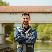 Person mit blauer Jacke steht vor einem Gewässer mit Brücke und rotem Dach im Hintergrund.