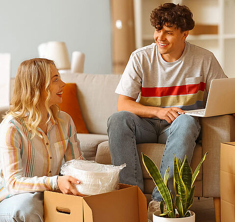 Zwei Personen sitzen zwischen Umzugskartons in einem Wohnzimmer mit Couch, Regal und Pflanzen.