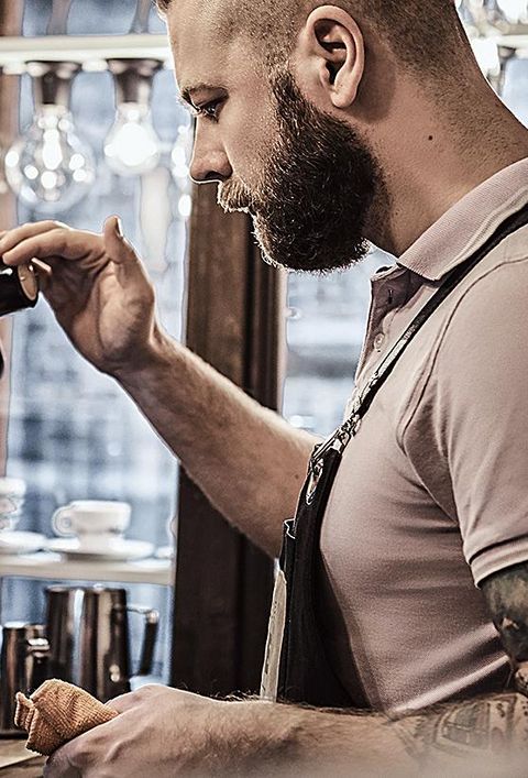 Barista in Uniform, der eine Tasse Kaffee für einen Kunden im Café zubereitet. Dampfende Kaffeemaschine