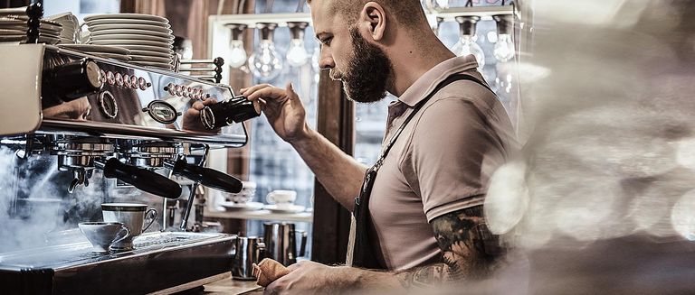 Barista in Uniform, der eine Tasse Kaffee für einen Kunden im Café zubereitet. Dampfende Kaffeemaschine