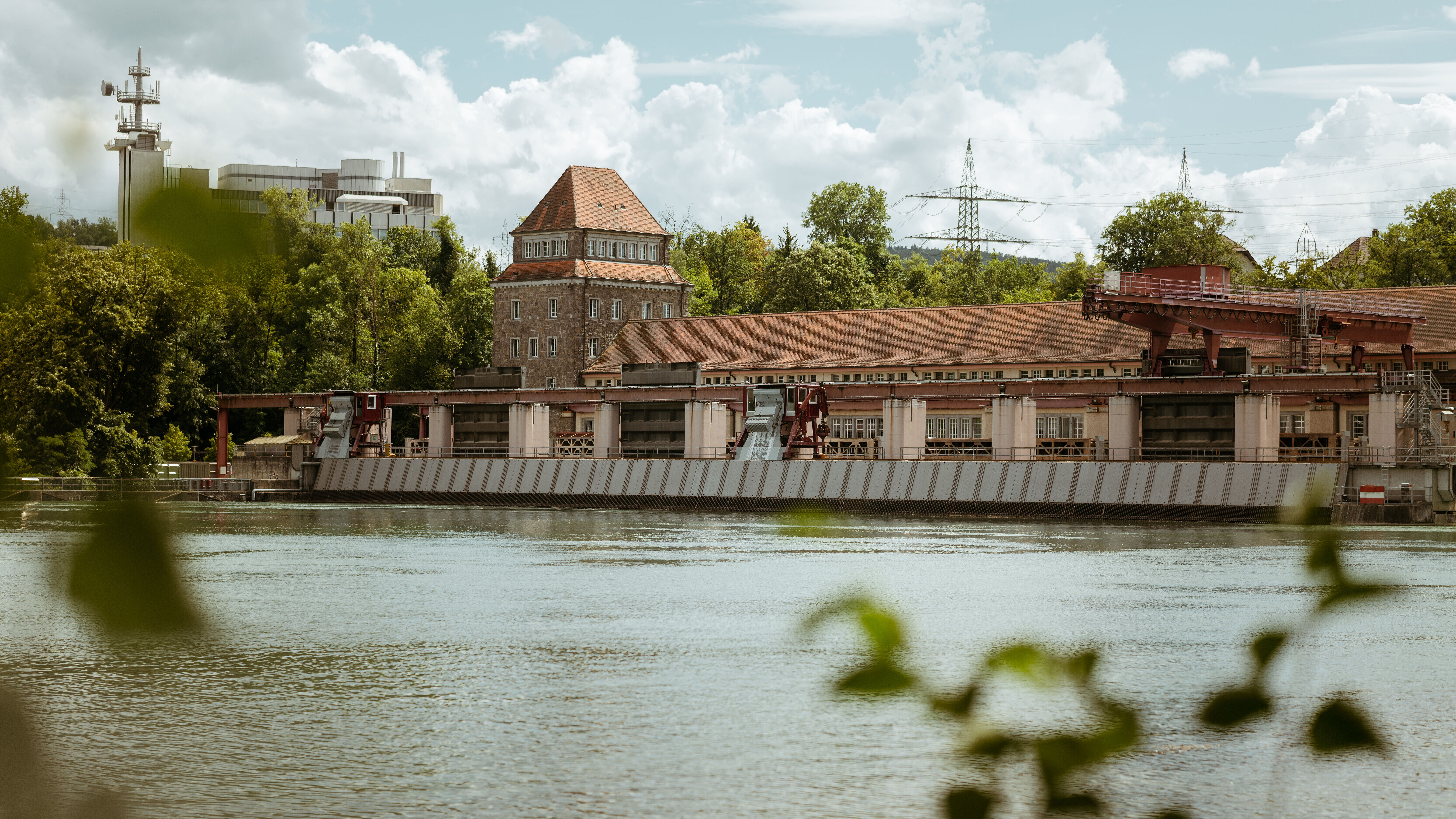 Wasserkraftwerk am Fluss mit Gebäuden, Wehranlage und Stromleitungen im Hintergrund.