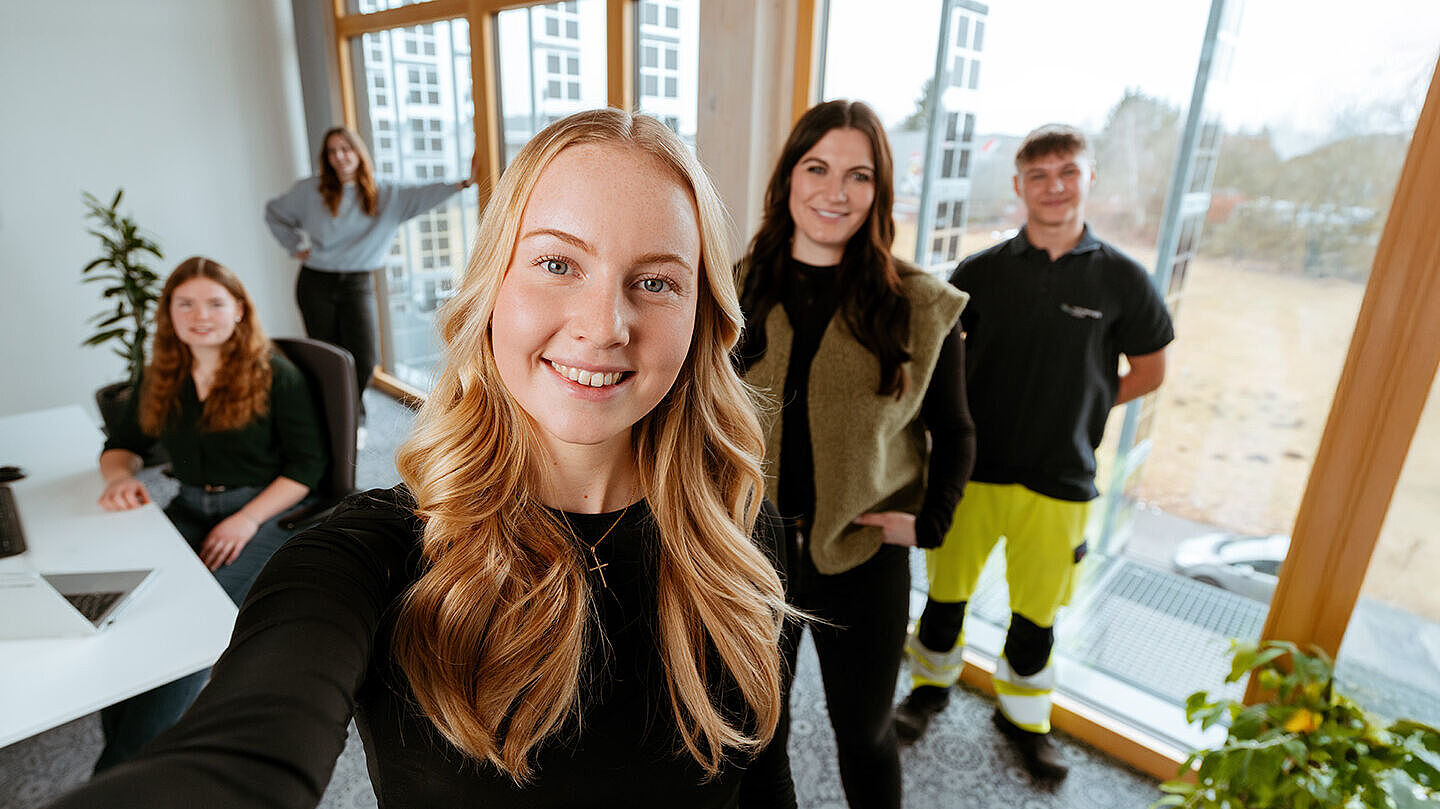 Mehrere Personen in einem Büro mit Schreibtisch, Fenstern und Arbeitsplätzen.