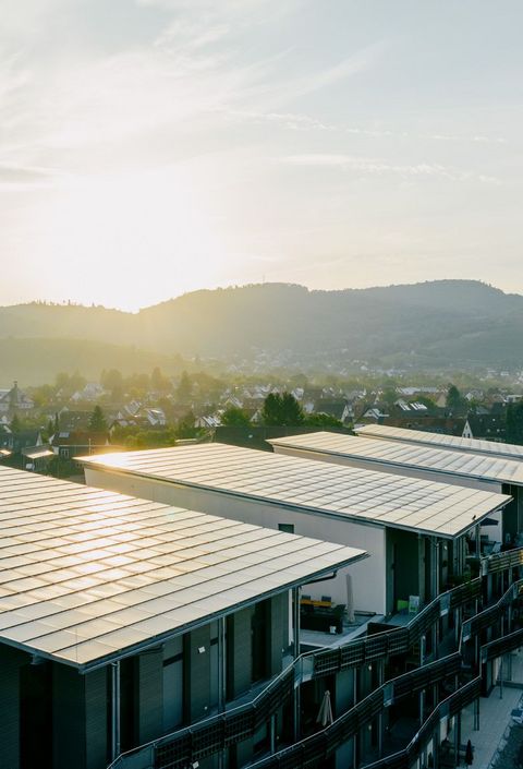 Solaranlage von naturenergie in Schallstadt auf den Dächern von Gebäuden in einer Wohngegend. Im Hintergrund sind Hügel und eine Kirche unter einem bewölkten Himmel bei Sonnenuntergang zu sehen.
