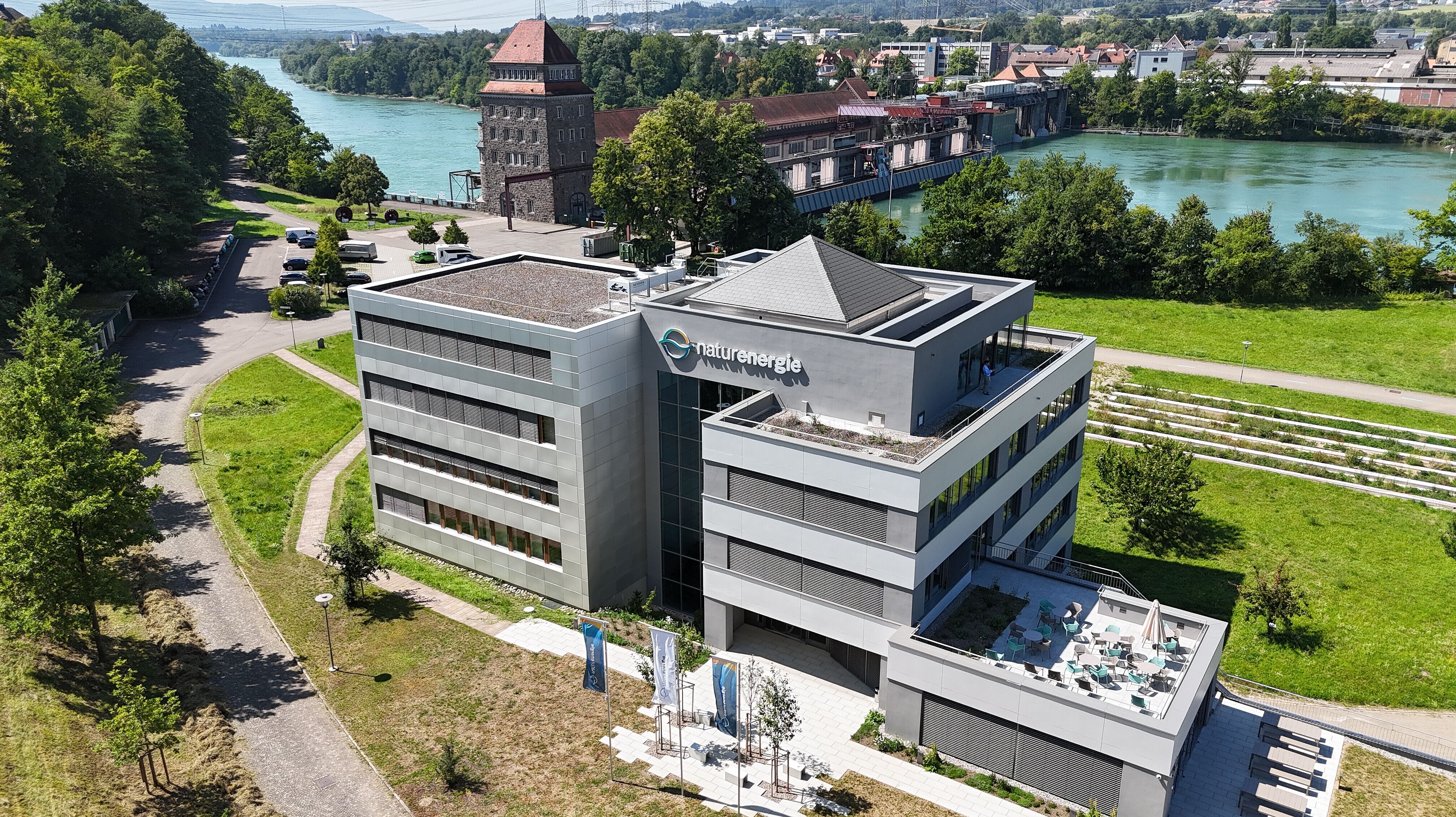 Luftaufnahme eines modernen Bürogebäudes der naturenergie direkt an einem Fluss. Hinter dem Gebäude befindet sich eine Wasserkraftanlage, umgeben von Bäumen, Wegen und einer grünen Landschaft.