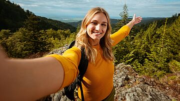 Person mit Rucksack macht Selfie auf Felsvorsprung in bewaldeter Landschaft
